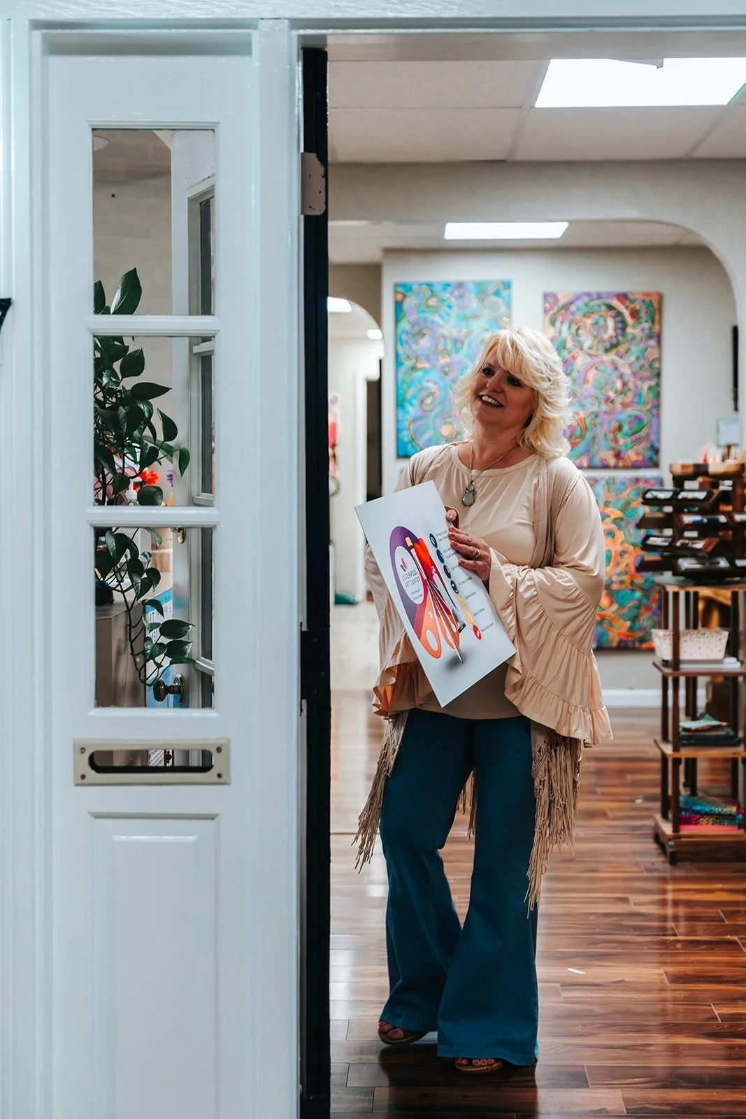 A woman with blonde hair smiling and holding a colorful brochure, standing in an art-filled room with paintings in the background.