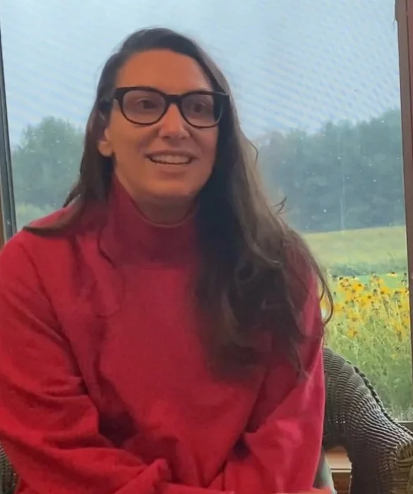 A woman with long brown hair and glasses, wearing a red sweater, sitting indoors near a window with a view of a field and trees.