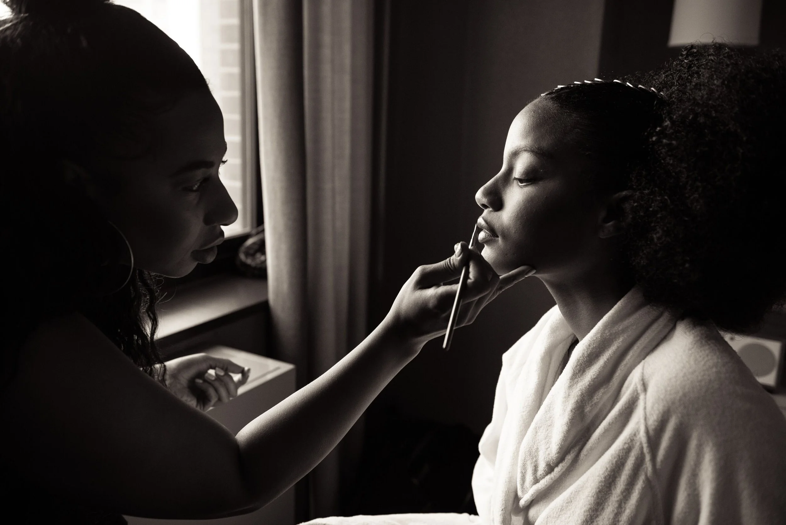 Two women are in a close, intimate moment. One woman is applying makeup or lip gloss to the other woman's lips. The scene is set indoors with a window and curtains in the background. The image is in black and white.