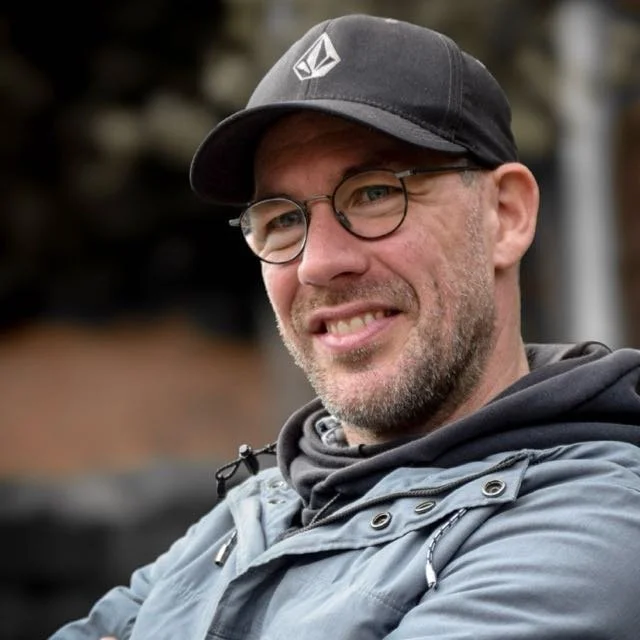 Micah Purnell wearing glasses, a black cap, and a pale blue jacket outdoors.