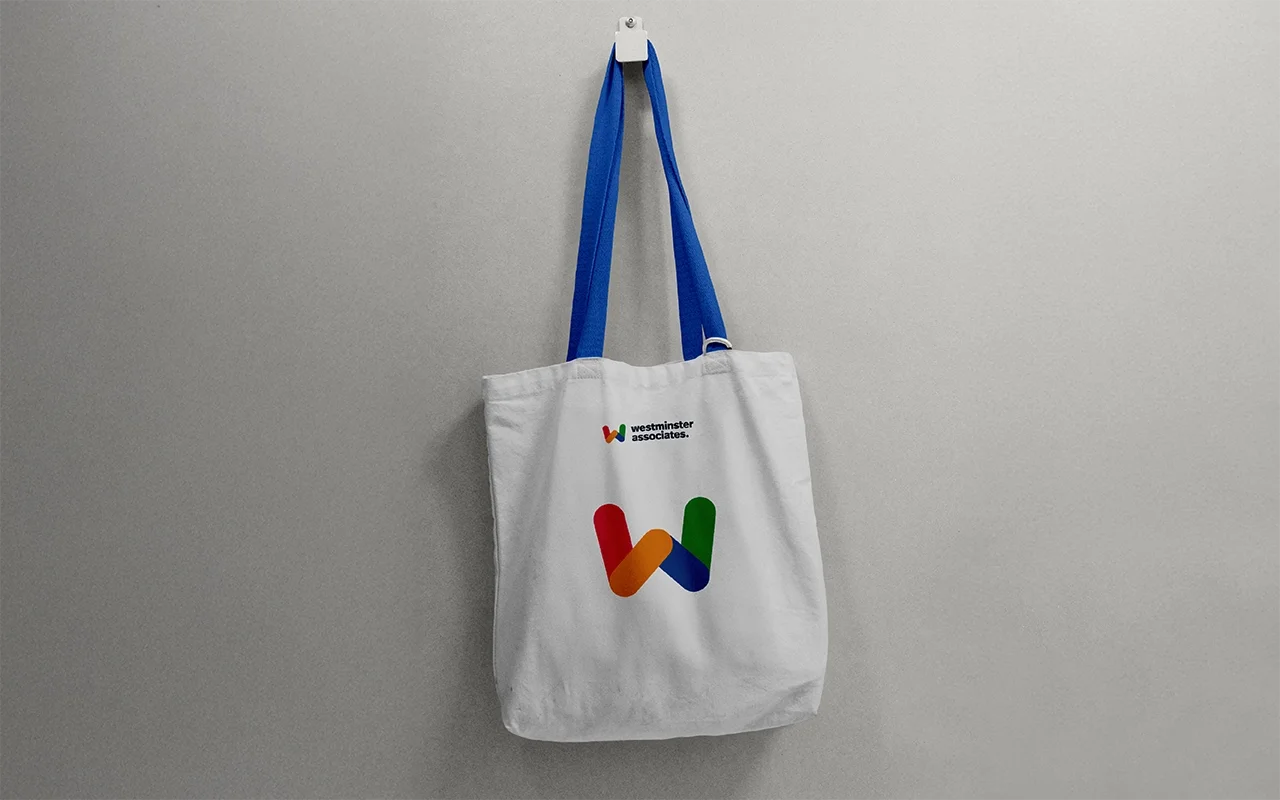A white tote bag with a multicolored 'W' logo and the text "westminster associates" hanging from a blue strap on a white wall.