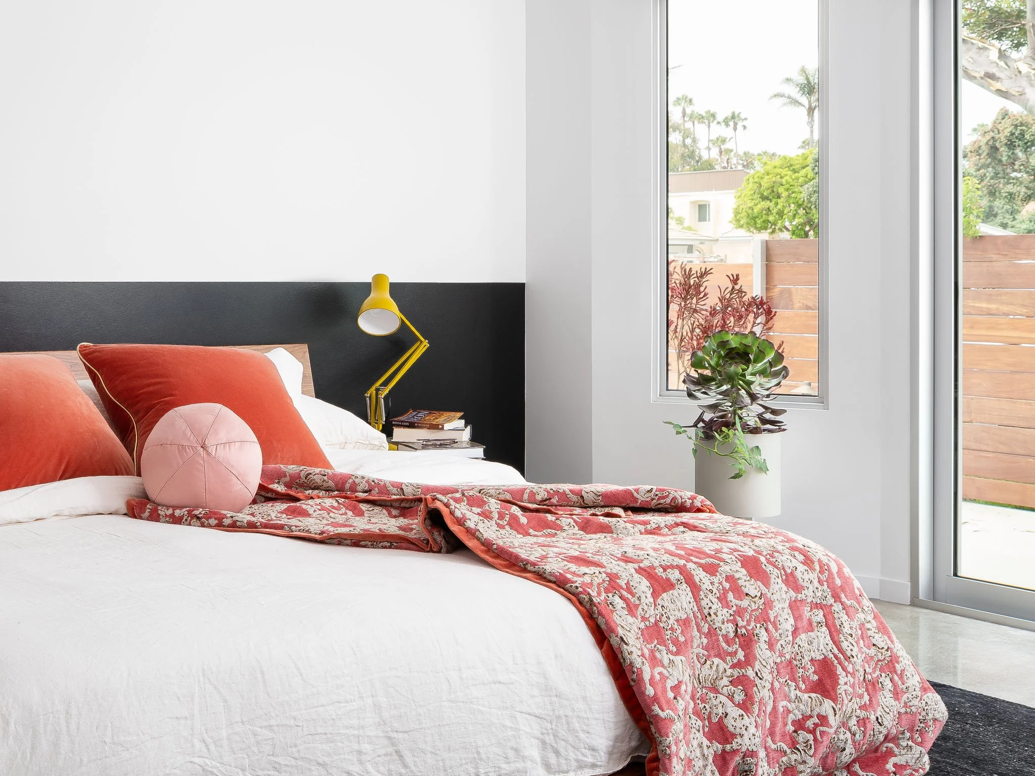 Modern bedroom at Channel Islands Harbor in Oxnard, California with white walls, a black accent wall, red and white bedding, a yellow lamp, and large windows.