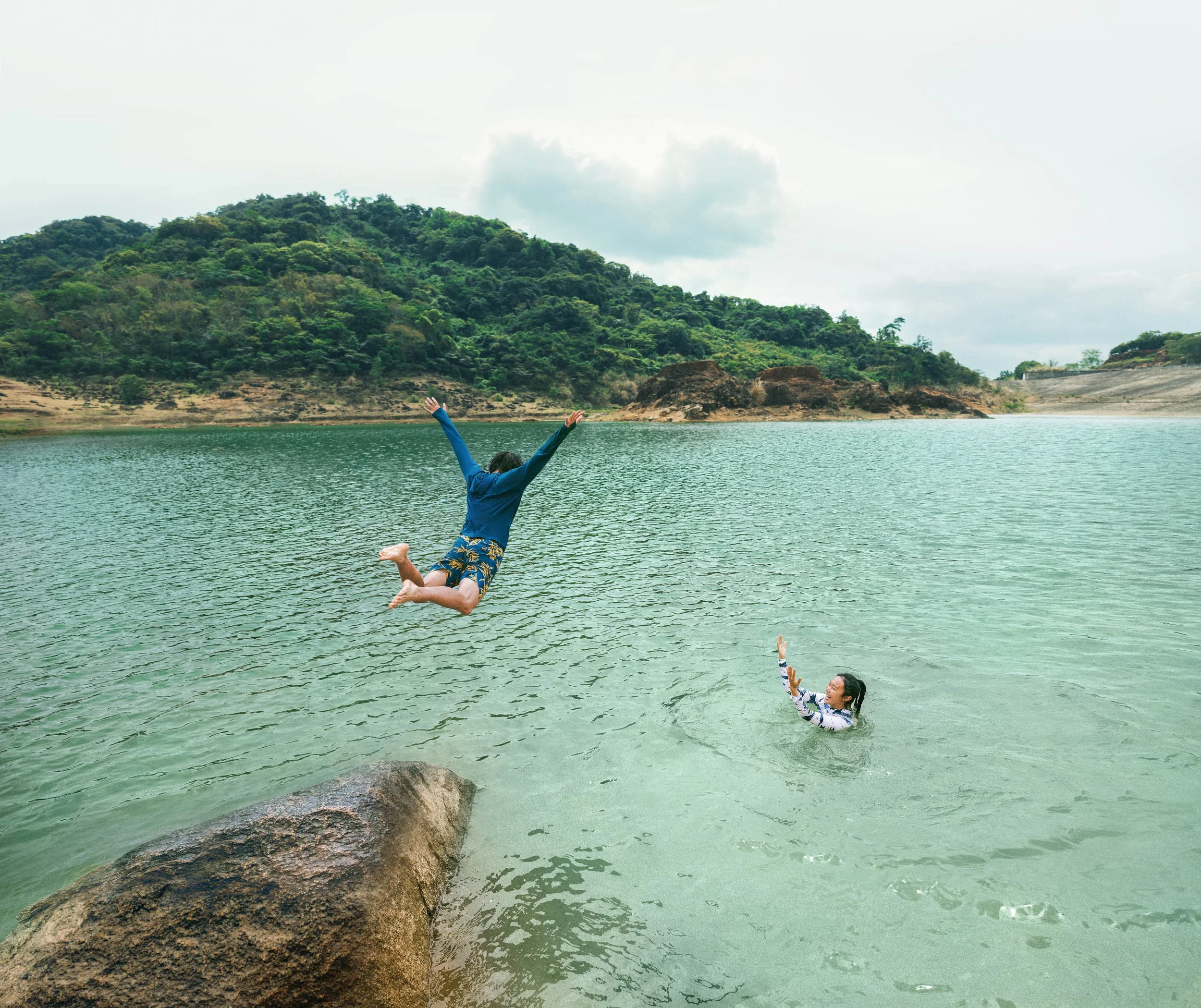 The North Face campaign - Young couple playing in a lake, one jumping off a rock and the other swimming with her hand raised, surrounded by green hills and cloudy sky photographed by Tang Ting in Hainan.