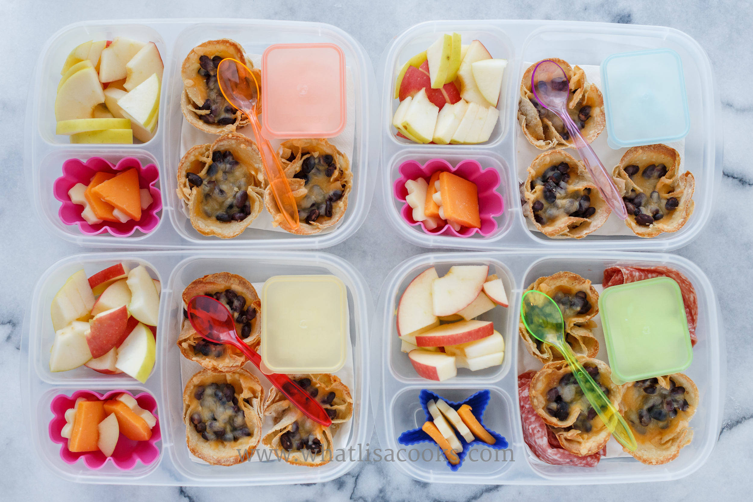 Corn tortilla cups with shredded cheese and black beans, cheese, apples, sour cream on the side, and one has a few slices of salami. 