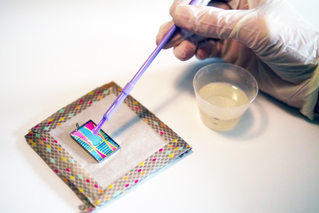 "Paint" in the resin. Be careful not to over fill the bezel cup. For this tutorial I put parchment paper on a piece of cardboard, to create a backer for the pendant so I could pour the resin on a flat surface. I taped the parchment paper down with washi tape. This is helpful if the eye of the pendant is large than the cup. You can push the eye into the cardboard so the pendant lays flat.