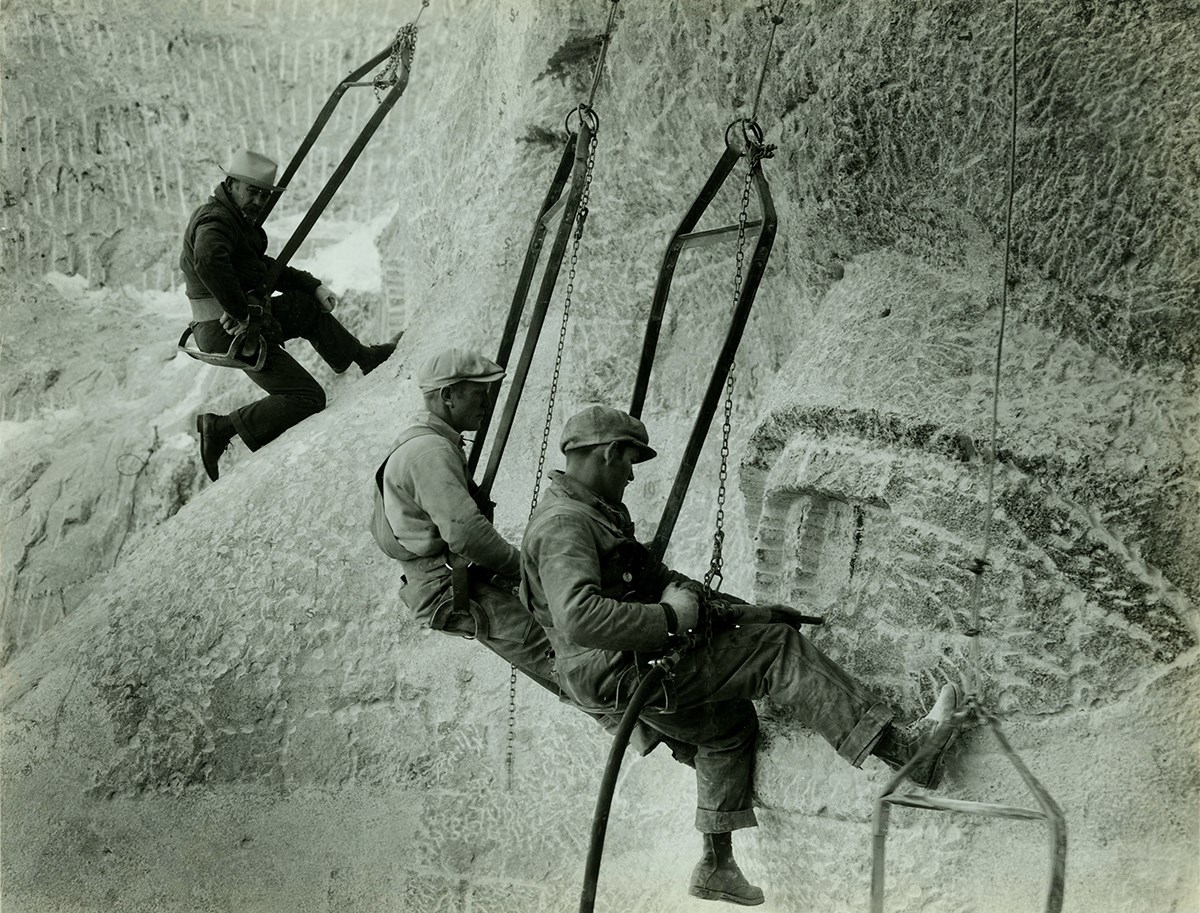 Carvers in Bosun's Chairs, jackhammering on Mount Rushmore, as Gutzon Borglum looks on.