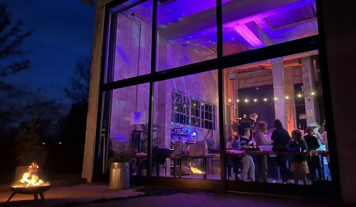 People gathered inside a rustic venue with wood and brick walls, colorful lighting, and string lights overhead, causing a lively and festive atmosphere.