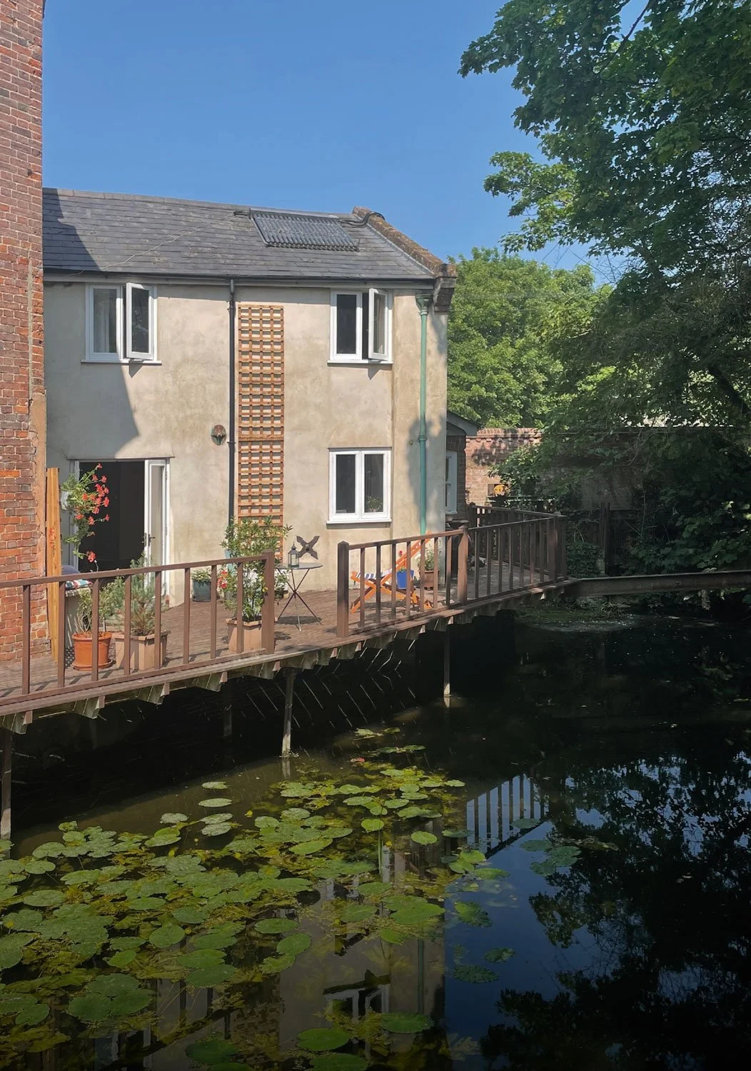A two-story house with a beige exterior, black roof, and white-framed windows, situated by a pond with lily pads, featuring a wooden deck with potted plants and outdoor furniture, surrounded by trees under a clear blue sky.