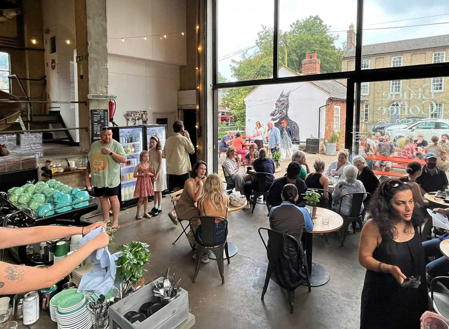 Inside a busy restaurant with people dining and socializing, large windows showing outdoor seating with more people, a mural of a dragon on the outside wall, and various tables and chairs inside.