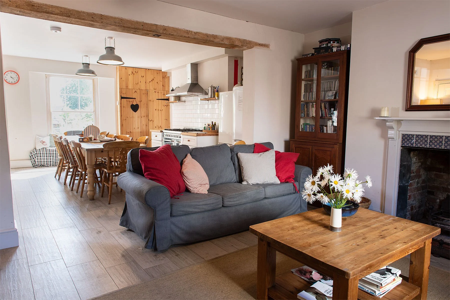 Living room and kitchen area with a gray sofa adorned with red and patterned cushions, a wooden coffee table with a vase of daisies, in front of a fireplace, with a wooden display cabinet and a dining table with chairs, and a window allowing natural light.