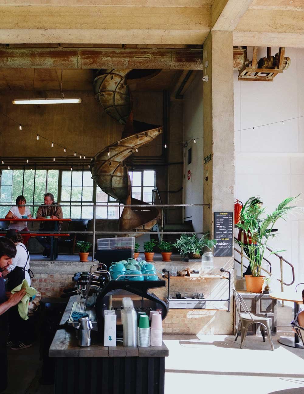 Interior of a cafe with a spiral slide in the background, hanging string lights, potted plants, and people sitting and working.
