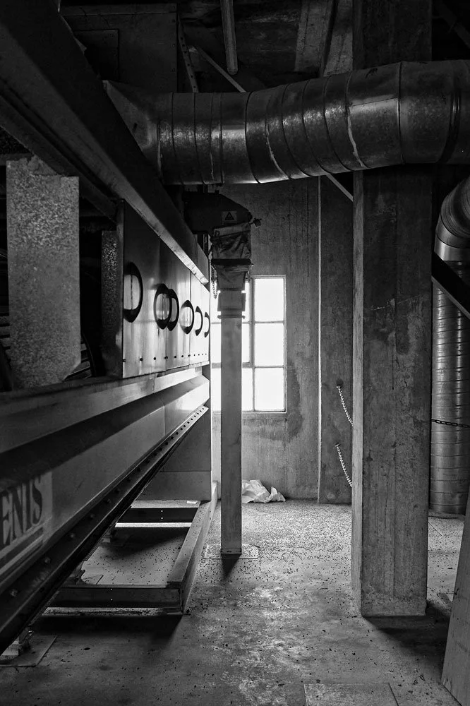 Black and white photo of an industrial interior with large ventilation ducts, a window allowing natural light, and machinery or equipment along the wall.