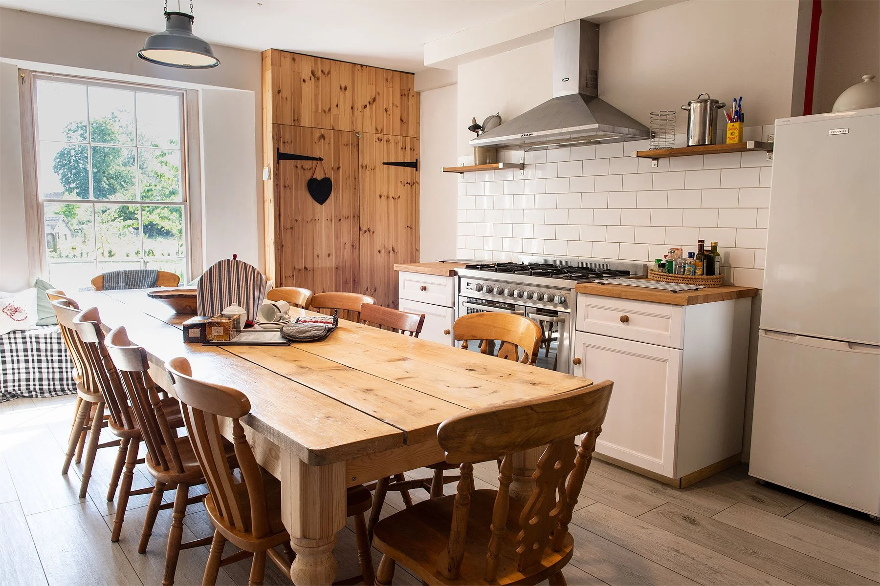 A cozy kitchen with a wooden dining table surrounded by wooden chairs, a large window letting in natural light, and white cabinets with a stainless steel stove and vent hood. There are open shelves with kitchen items and a white refrigerator on the right.