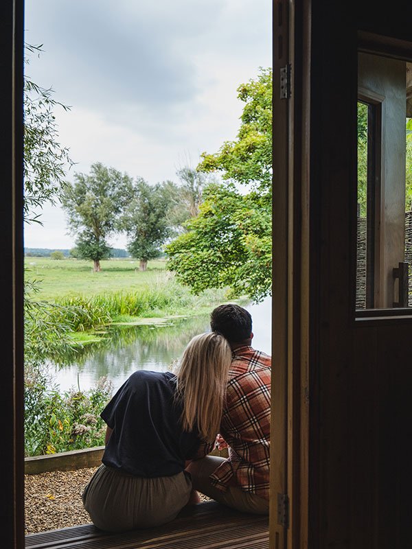 A woman and a man sit close together on a porch, looking out at a peaceful river with trees in the background.
