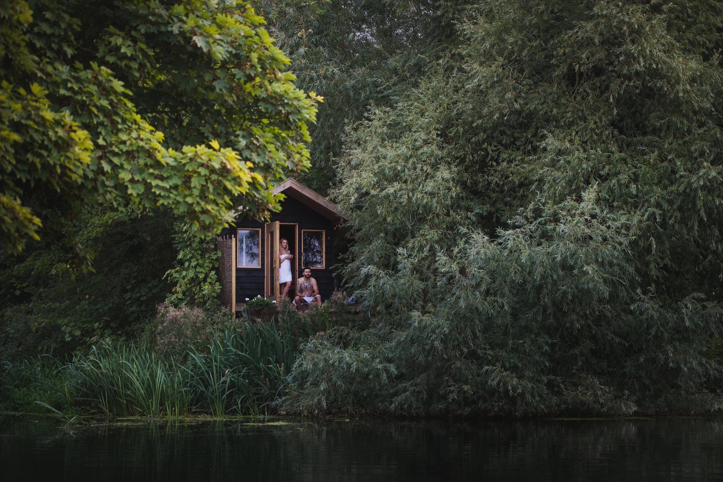 A small black wooden house on a riverbank, surrounded by dense green trees and bushes, with two people standing and sitting on the porch.