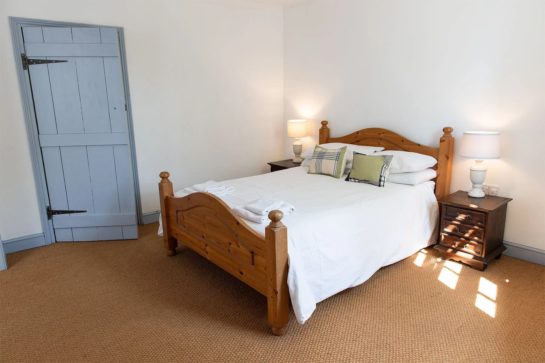 A neatly made bed with white linens and decorative pillows, flanked by two wooden nightstands with lamps, in a cozy bedroom with beige carpet and a closed light blue door.