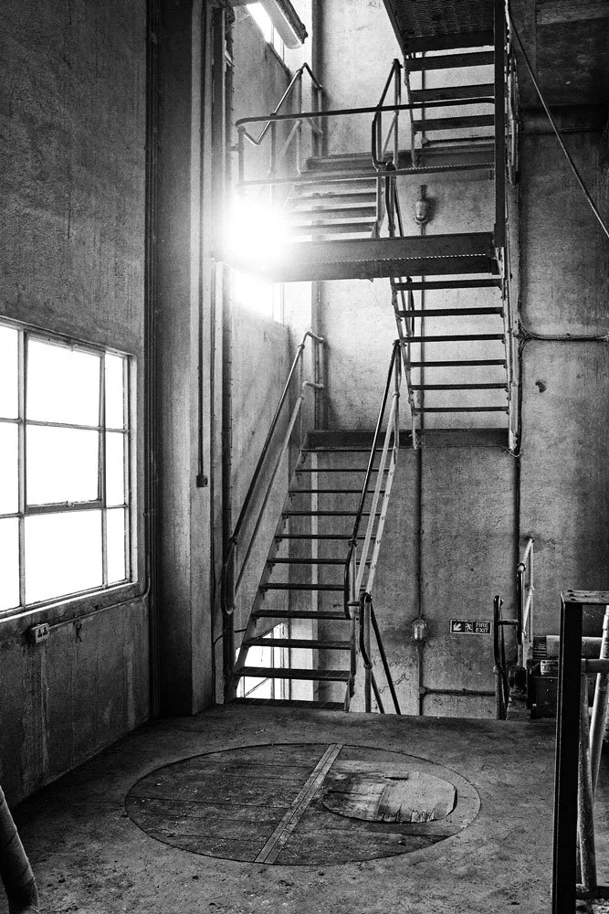 Industrial staircase with metal railings in a concrete building, sunlight shining through a window, and a round hatch on the concrete floor.
