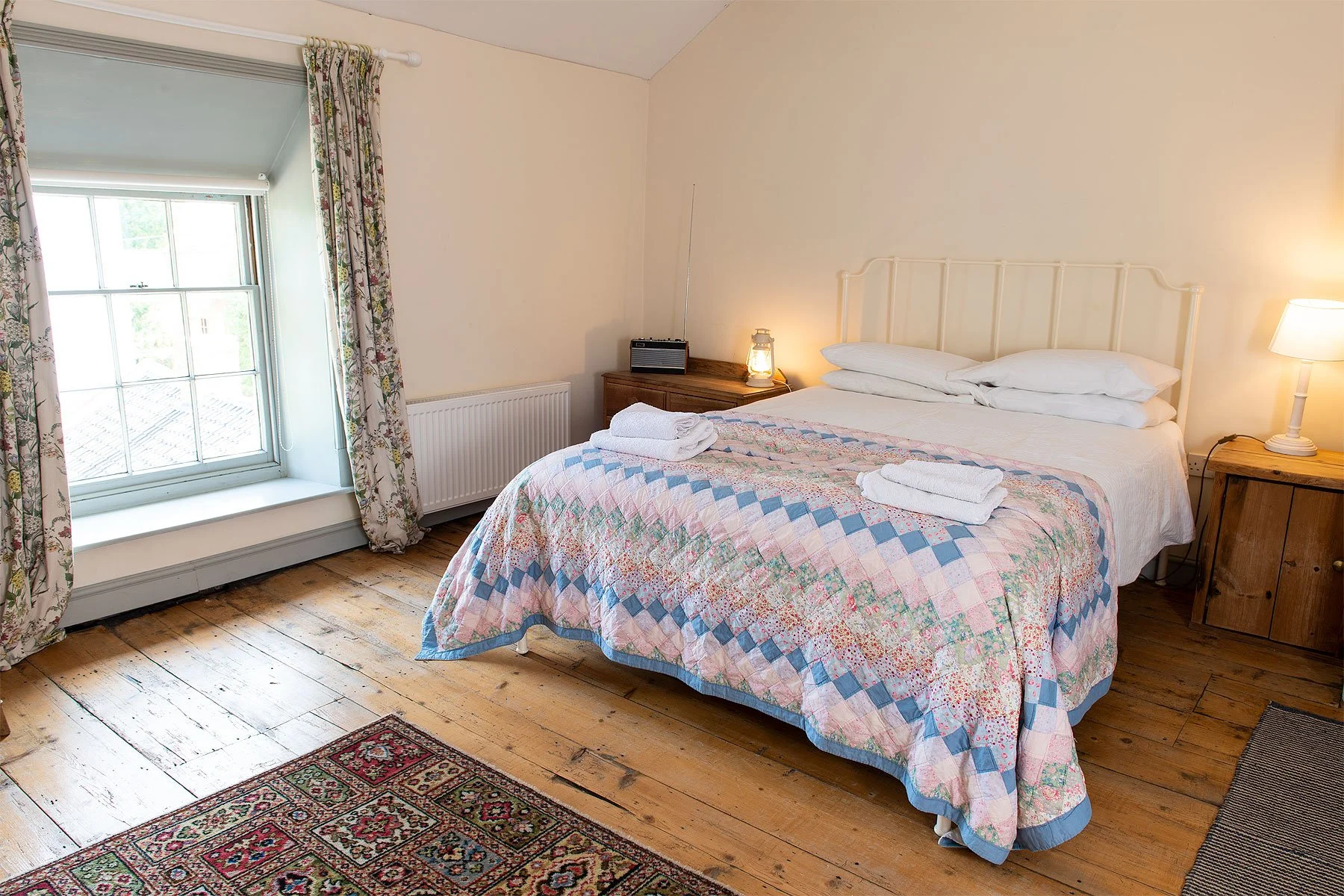 A cozy bedroom with a large bed covered in a pastel quilt with pink, blue, and green patterns, and two rolled towels on top. There are two wooden nightstands with lamps, a radio, and a candle. The room has wooden floors, floral curtains, and a window allowing natural light.
