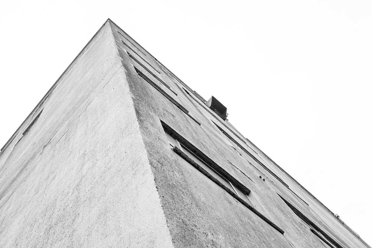 Black and white photograph of the corner of a tall, textured building taken from a low angle, showing windows and a clear sky.