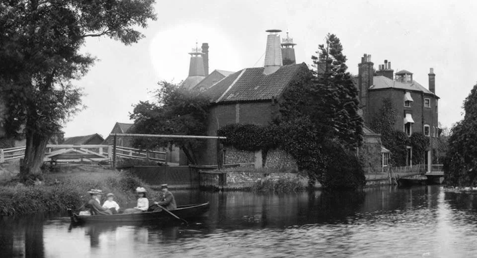 A black and white photo showing a small boat with four people on a river next to a large, old house with a chimney, surrounded by trees and a fence.