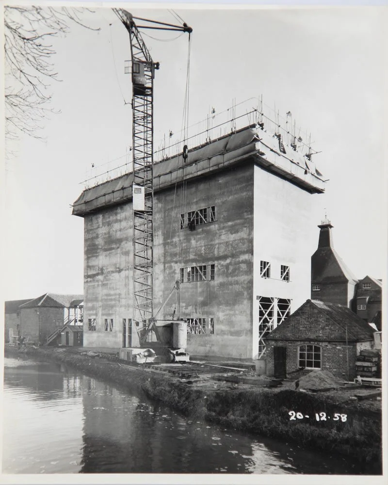 Construction site of a large concrete building next to a water body, with a tall crane and scaffolding. The building is still under construction.