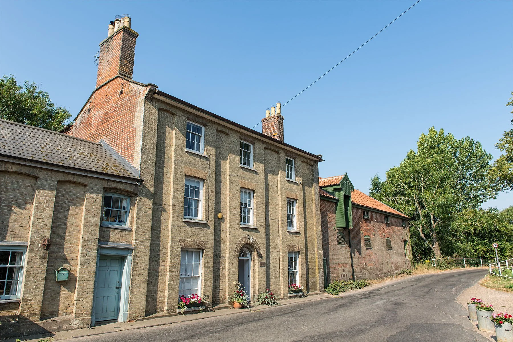 Historic brick building on a curved street with trees and flower pots, under a clear blue sky.
