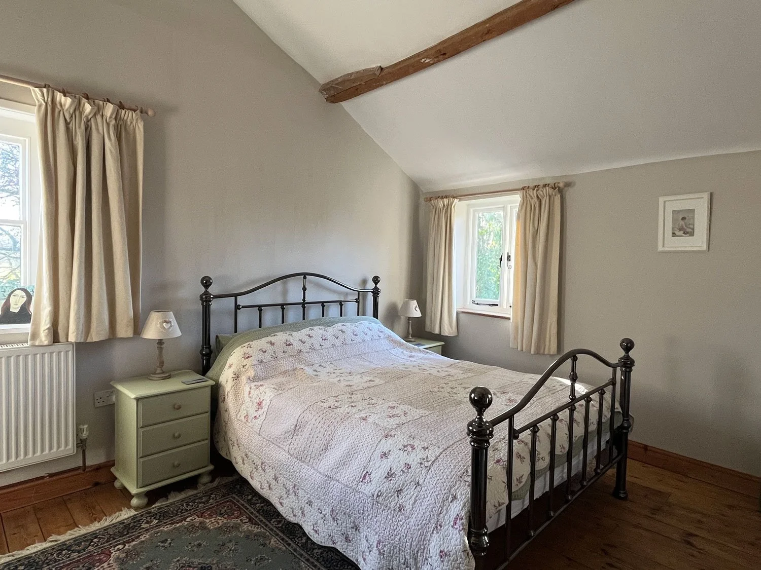 A cozy bedroom with a metal bed frame, a floral quilt, two bedside tables with lamps, and windows with cream curtains.