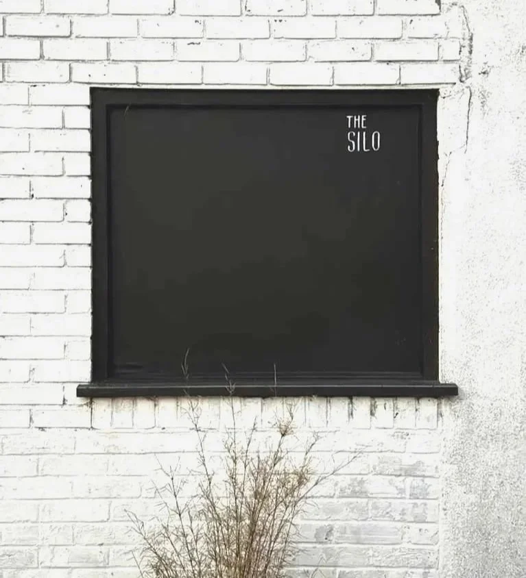A black chalkboard with the words 'The Silo' written in small white letters in the top right corner, mounted on a white brick wall with a small dry plant at the base.