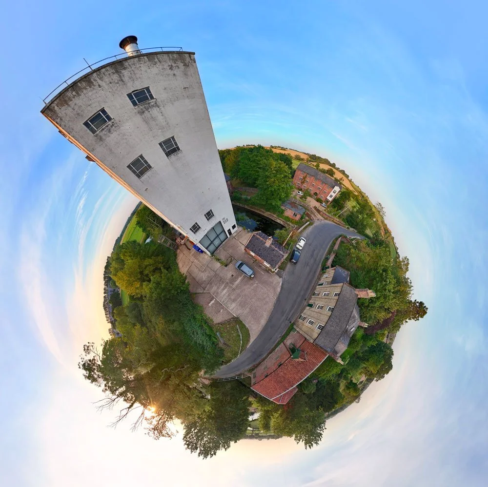 A fisheye photograph of a small town showing a tall white building, trees, houses, a road, and a clear sky with the sun setting.