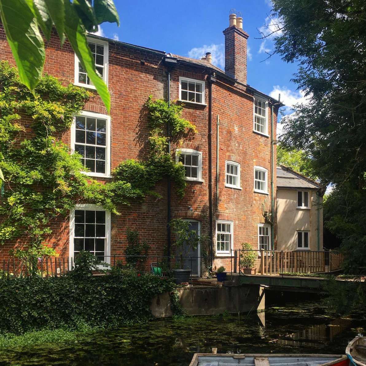 A multi-story brick house with white-framed windows, surrounded by trees and greenery. There is a small waterway or canal in the foreground with a boat docked, and a wooden deck with plants on it.