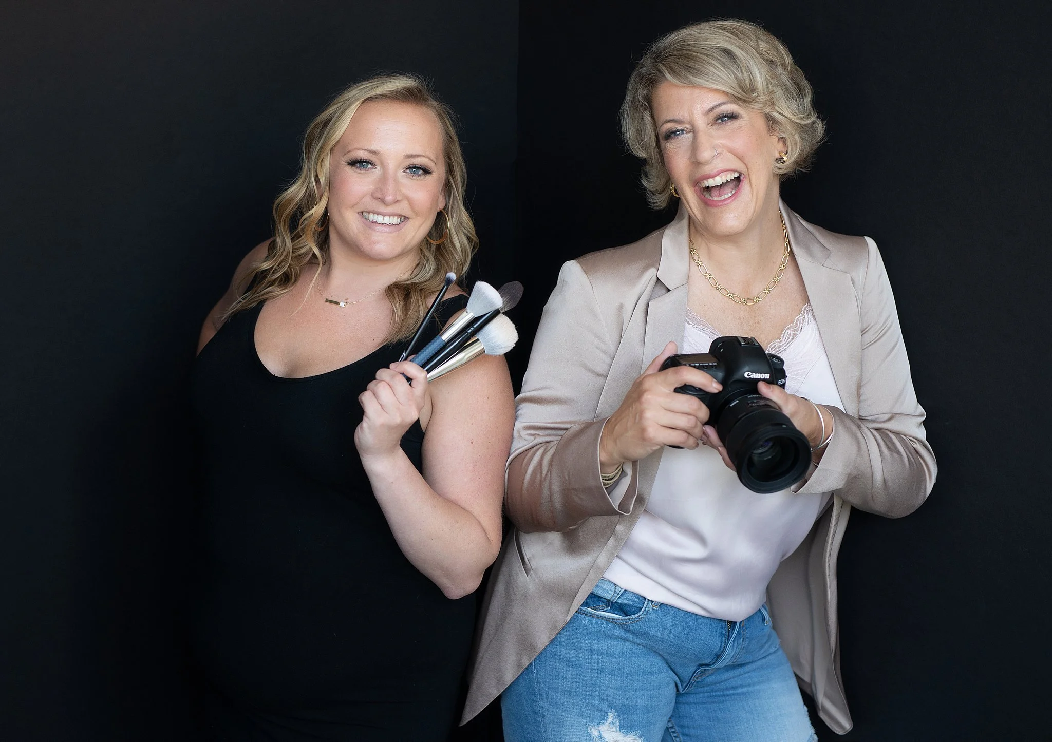 Two women standing against a black background, one holding makeup brushes and the other holding a Canon camera, both smiling and appearing happy.