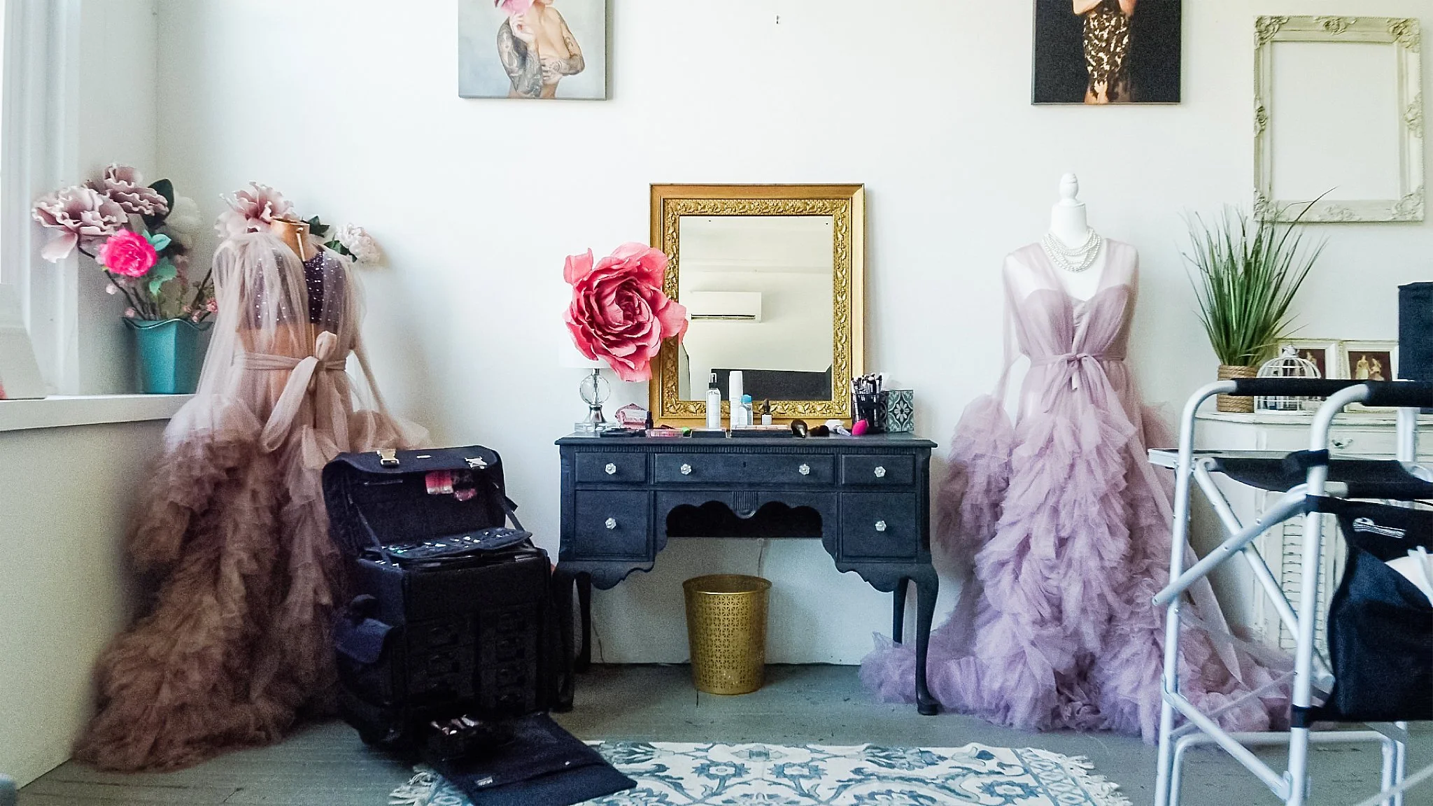 A dressing room with two mannequins: one on the left wearing a pink gown with a veil and the other on the right wearing a lavender gown, positioned in front of a black vanity table with a mirror, makeup, and products. There are flower decorations on the wall and a potted plant on a dresser on the right.