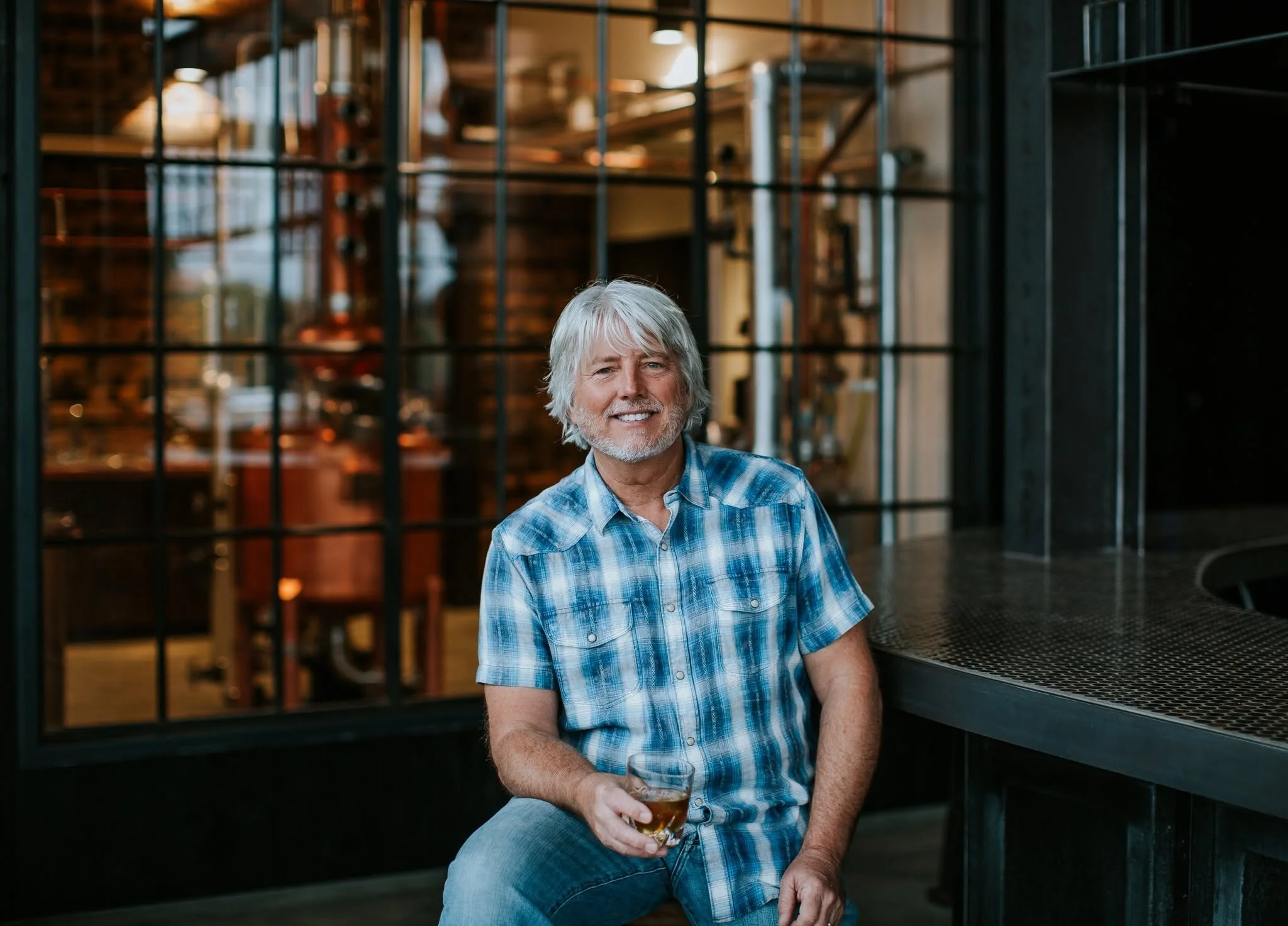 A smiling Wally Dant, distiller, wearing a plaid shirt and holding a rocks glass of bourbon.