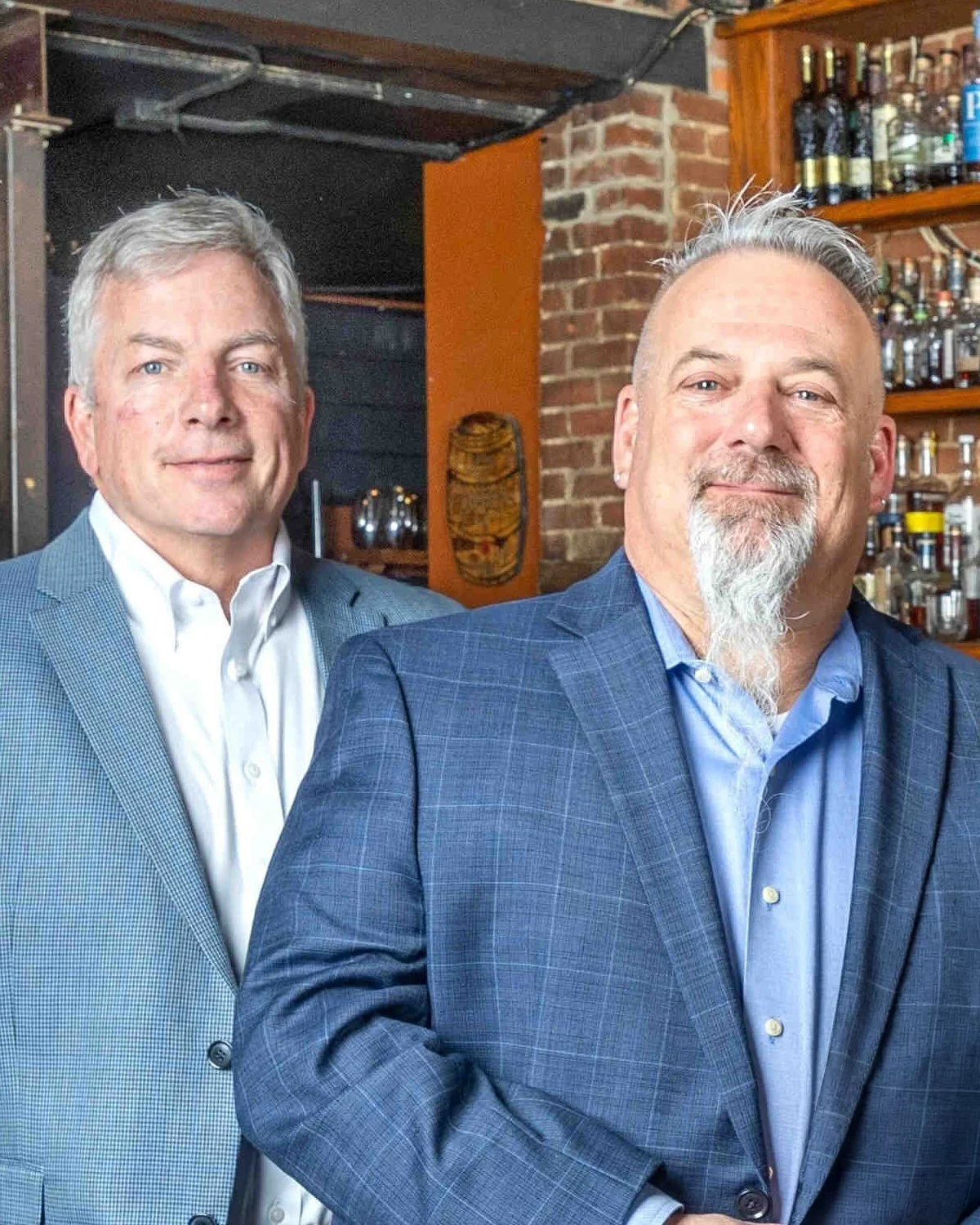 Two men in business suits standing side by side inside a bar or restaurant, with shelves of liquor bottles and brick wall in the background.