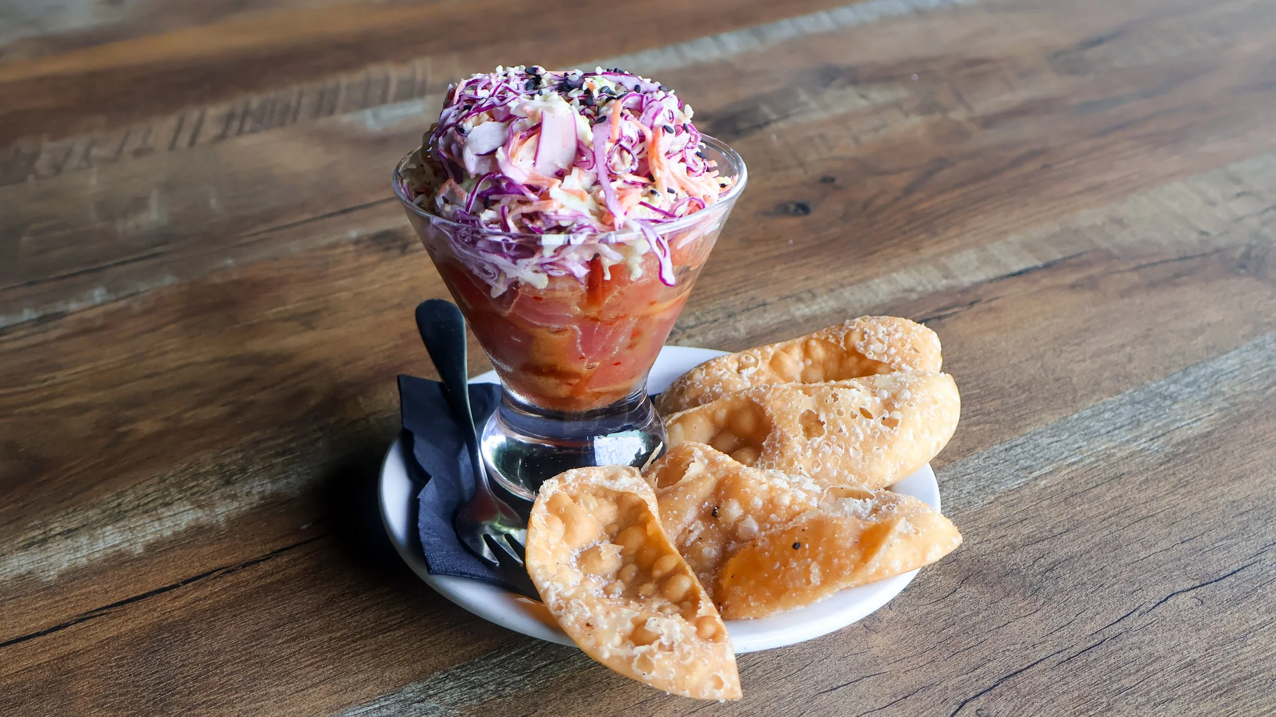 Tuna Poke served in a clear glass topped with a multi-colored cole slaw. Crispy gyoza chips on the side. Served on a white plate sitting on a wooden table.