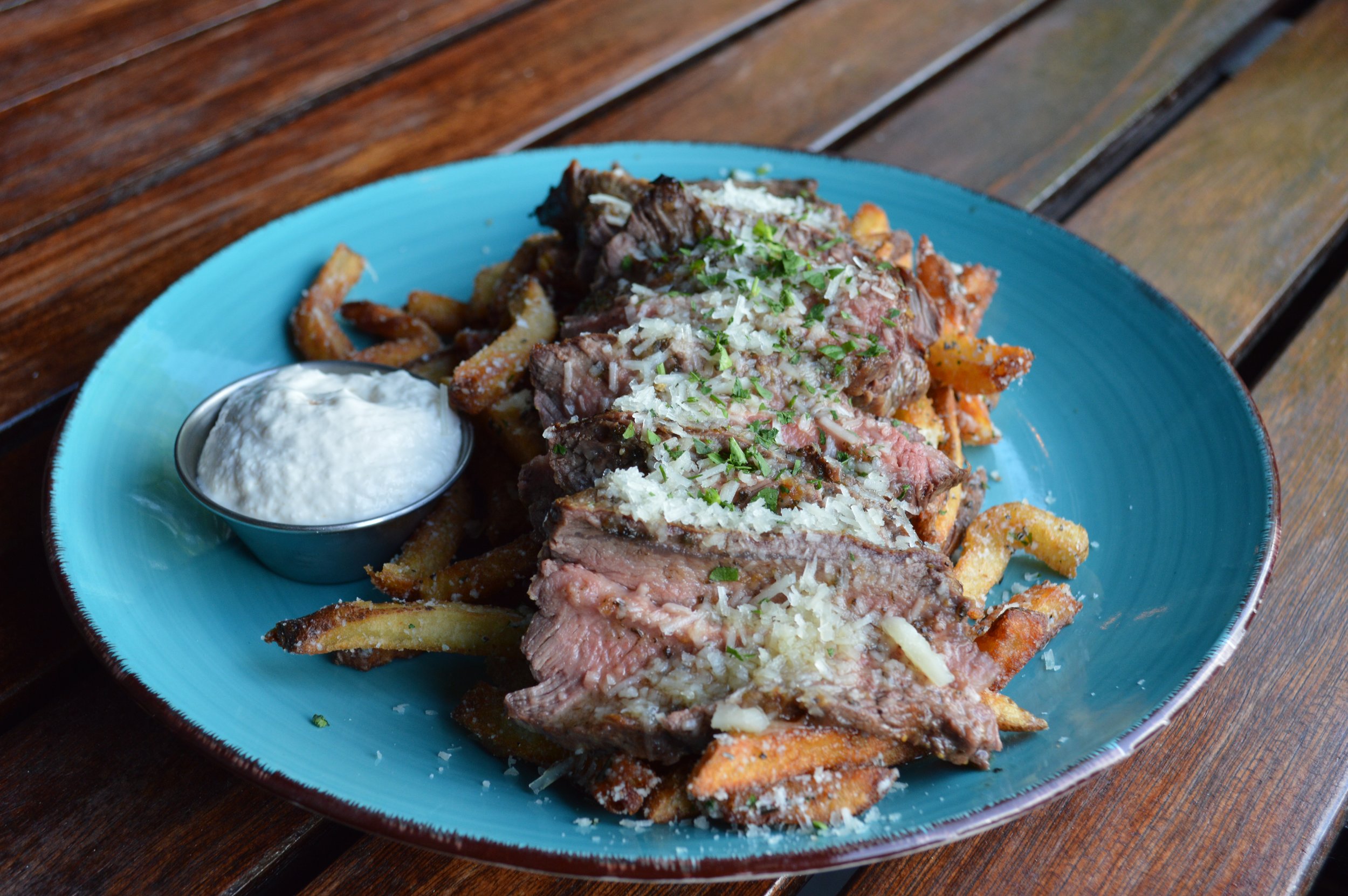 A round blue plate with a layer of seasoned french fries topped with sliced rare steak and parmesan cheese. A ramekin full of white horseradish sauce is on the side