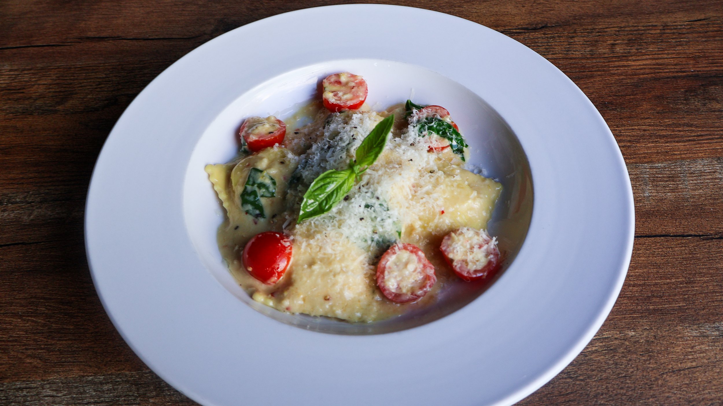 Ravioli with tomatoes, basil, and cheese served in a white bowl sitting on a wooden table.