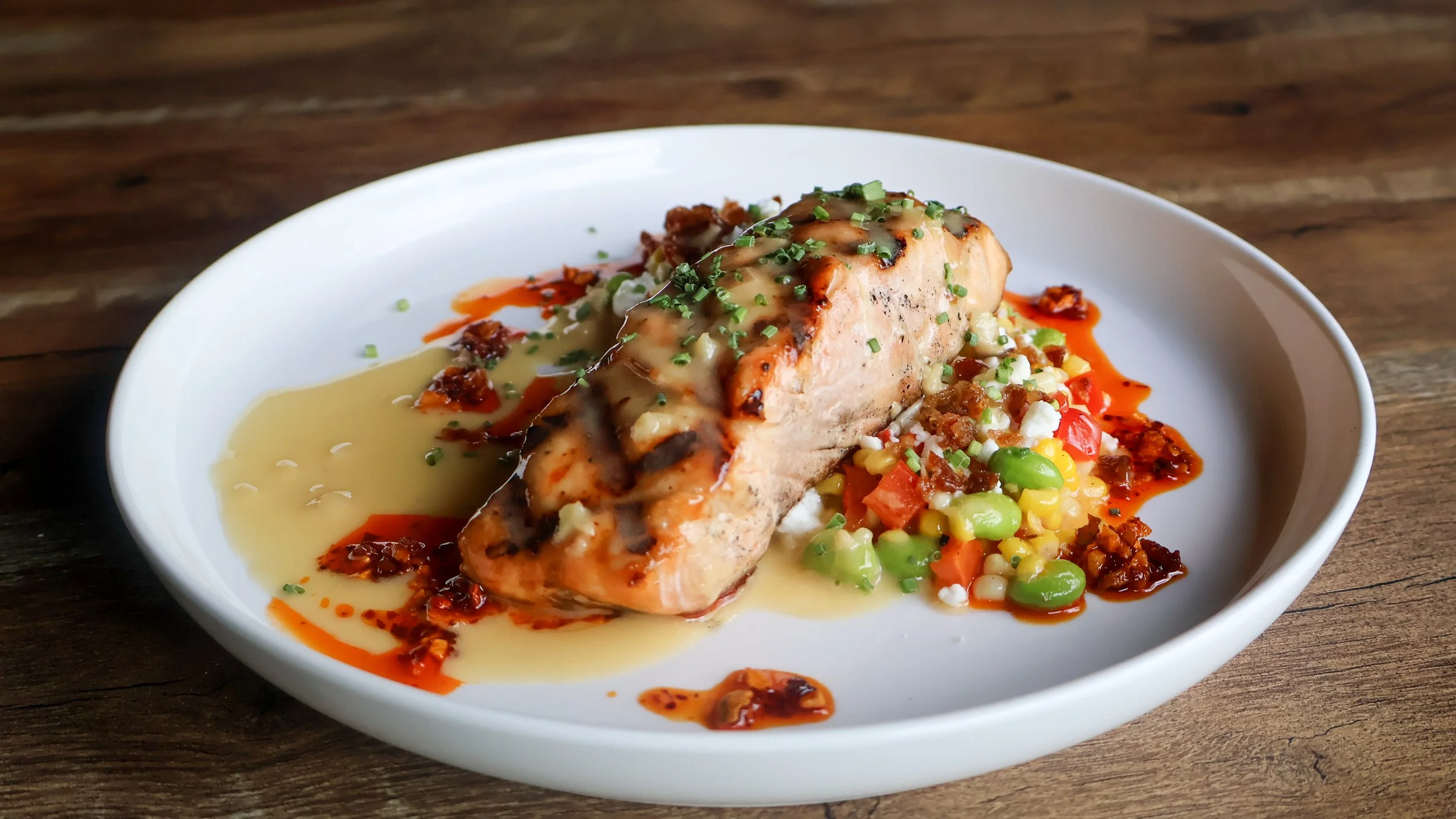 Grilled salmon filet with corn and edamame succotash, lemon sauce and chili oil on a white plate.