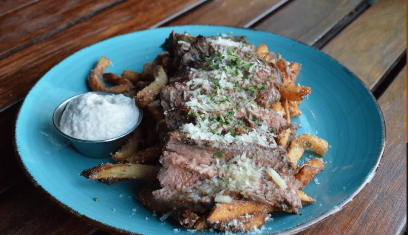 A round blue plate with a layer of seasoned french fries topped with sliced rare steak and parmesan cheese. A ramekin full of white horseradish sauce is on the side