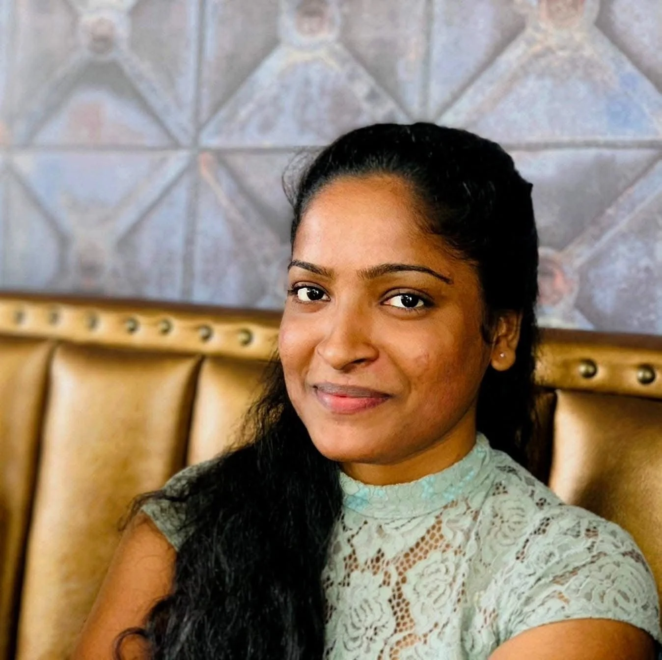 Apsara - genetics researcher. A woman with long dark hair and a grey lace top sitting on a tan-colored upholstered seat, with a textured wall in the background.