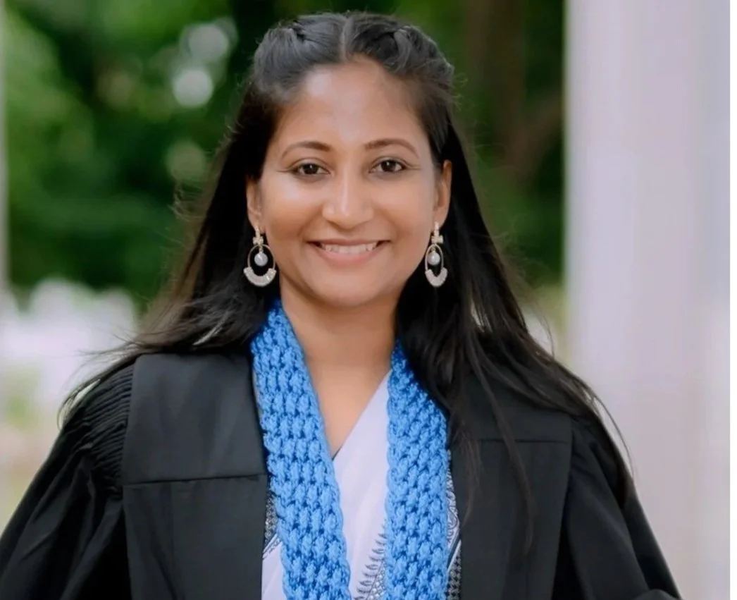 Malsha - joint project manager (policy). A woman in graduation attire with a black gown, white clothes, and a blue stole, smiling outdoors with blurred greenery in the background.