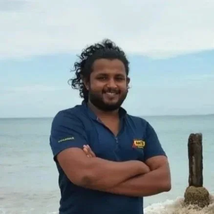 Neranjan - senior researcher. A man with curly hair and a beard standing on a beach with arms crossed, smiling at the camera. The ocean and sky are in the background.