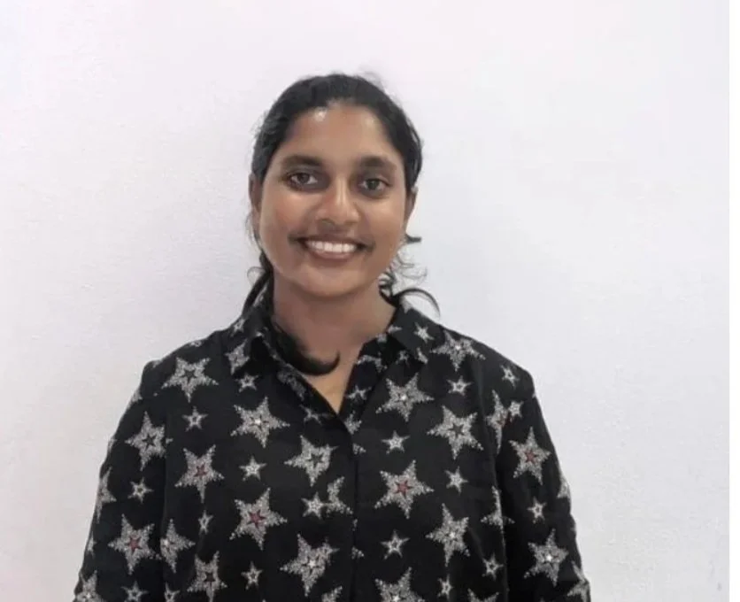 Chethana - senior researcher. A woman with dark hair in a ponytail, smiling, wearing a black shirt with silver star patterns, standing against a plain white wall.