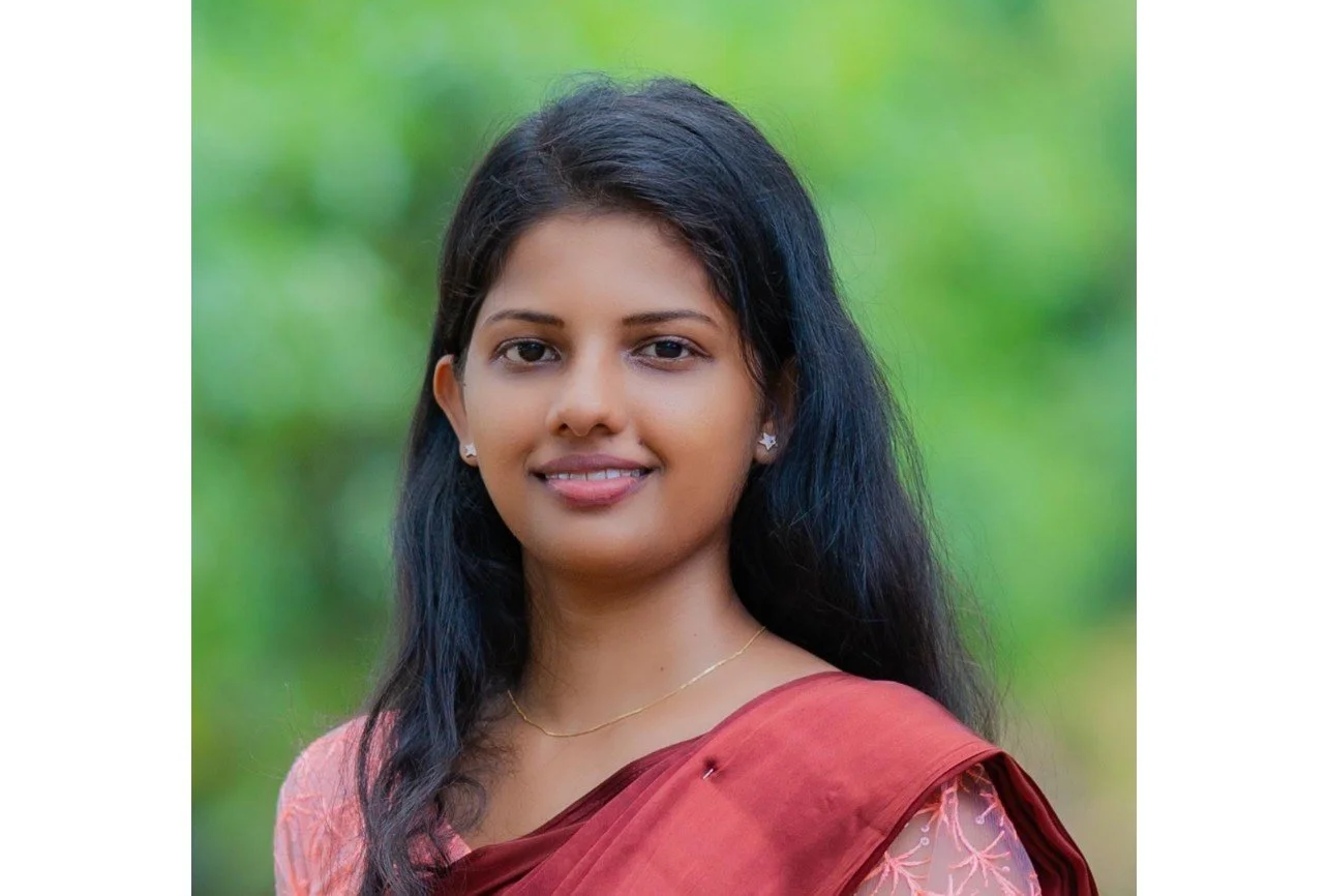 Dilrukshi - senior researcher. A young woman with long black hair smiling outdoors, wearing a maroon sari with pink embroidery, small earrings, and a delicate gold necklace, with a blurred green background.