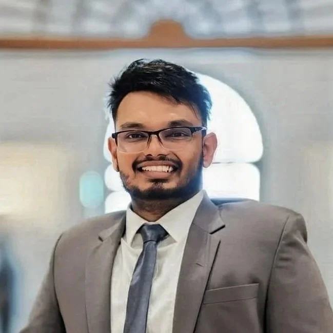 Henry - joint project manager (policy). A young man wearing glasses, a gray suit, and a tie, smiling, in an indoor setting with a blurred background.