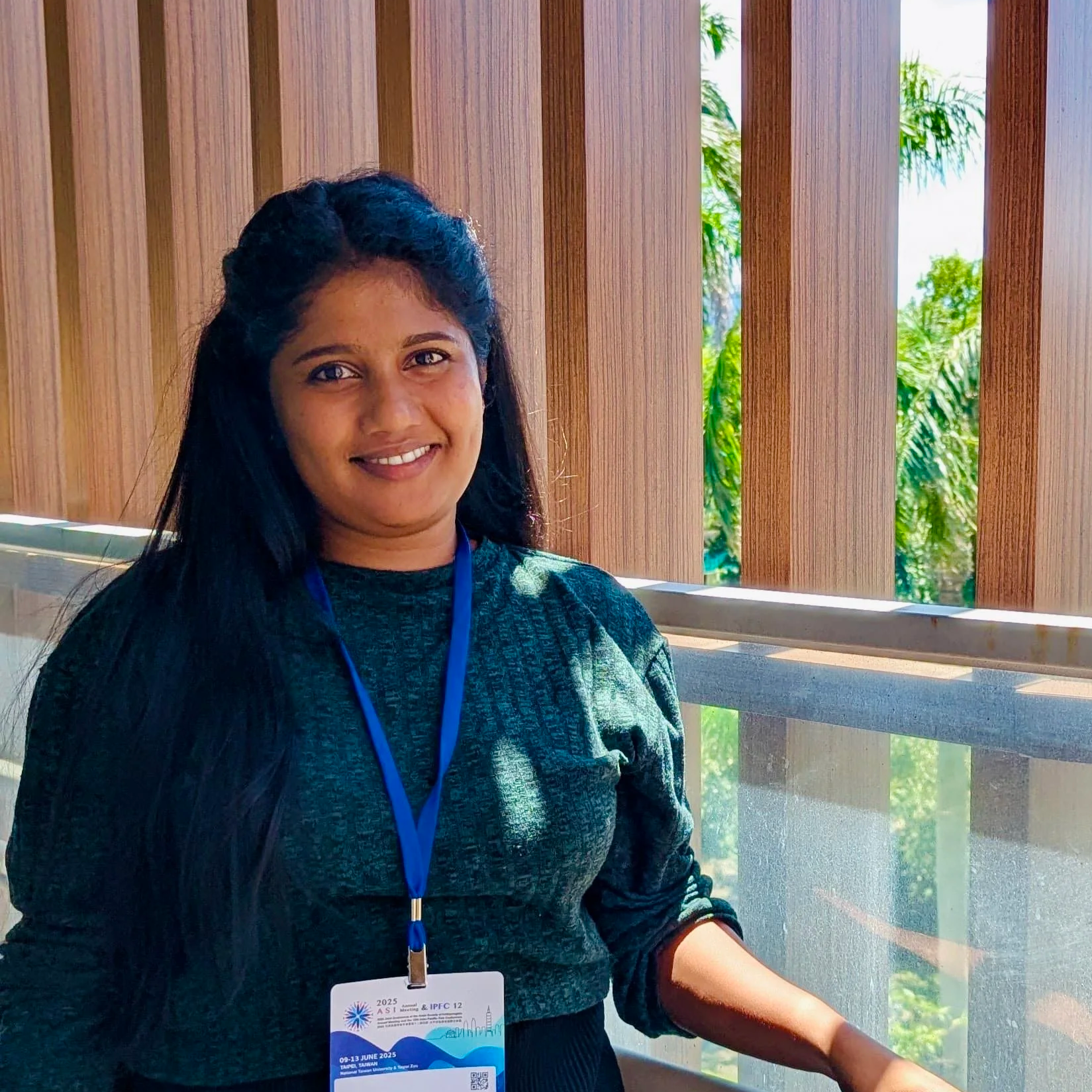 Maura - senior researcher. A woman with long black hair, wearing a dark gray top and a conference badge on a lanyard, smiling, standing indoors near a wooden wall with windows revealing green trees outside.