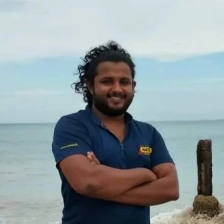 Neranjan - senior researcher. A man with curly hair and a beard standing on a beach with arms crossed, smiling at the camera. The ocean and sky are in the background.