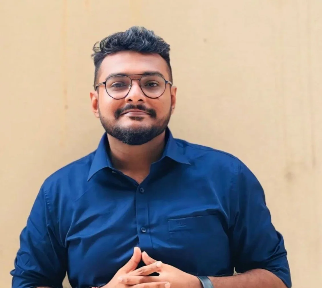 Shownath - senior researcher. A man with glasses, a beard, and dark hair wearing a blue shirt, standing against a beige background.