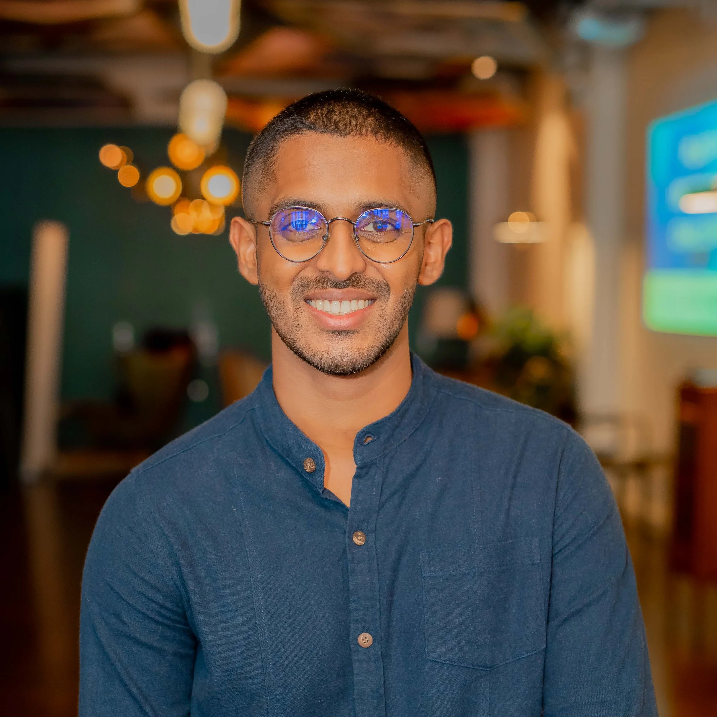 Rahal - fisheries programme manager. A smiling man wearing glasses and a blue button-up shirt in a warmly lit indoor setting.