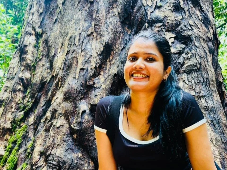 Pubudini - policy programme manager. A woman with long dark hair smiling outdoors next to a large tree trunk.